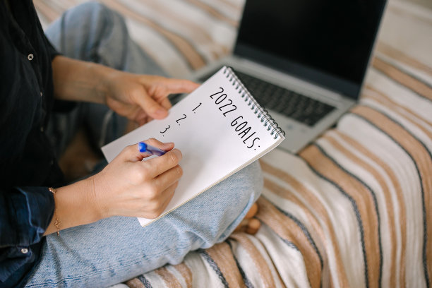 一名年轻女子一边在家用笔记本电脑在线工作，一边写下2022年的目标、任务清单和计划图片下载