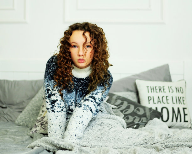 卷毛的女孩穿着一件暖暖的蓝白毛衣躺在床上，毯子是灰色的，她在枕头旁边的花瓶后面沉思和美丽的特写照片图片下载