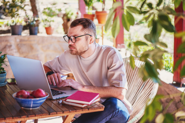 年轻迷人的男子，戴着眼镜，自由职业者在家工作，使用笔记本电脑，坐在露台上的一个乡村别墅的夏天图片下载