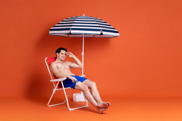 赤裸上身的白人男子穿着夏天的短裤，坐在躺椅上，打着沙滩伞，背景是彩色的演播室图片下载