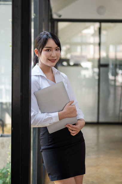 一名亚洲妇女抱着一台笔记本电脑站在玻璃墙前，照片上电脑盖是关着的。日常工作中使用的电子设备图片下载