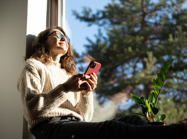 漂亮的卷发年轻快乐的女人，戴着眼镜，坐在窗台上俯瞰花园，手里拿着智能手机图片下载