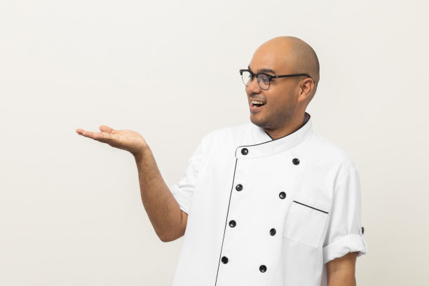 微笑的年轻英俊的亚洲男子厨师制服与眼镜站张贴显示手掌心举一些菜单上孤立。厨师职业厨师在厨房、餐厅和酒店工作。图片下载