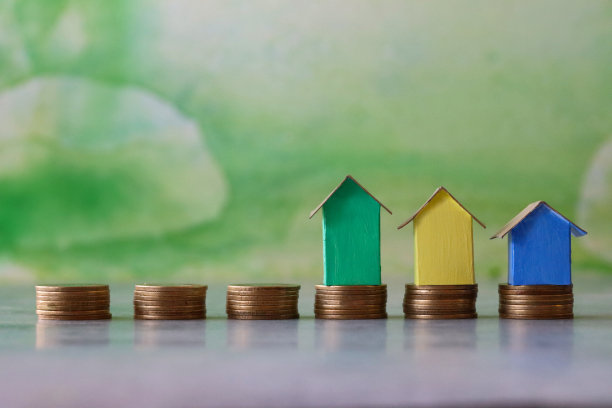 特写图像的一排排彩色纸板房子，五颜六色的海滩小屋站在成堆的硬币，重点前景，斑驳的绿色背景，家庭财务，房地产和假日储蓄概念图片下载