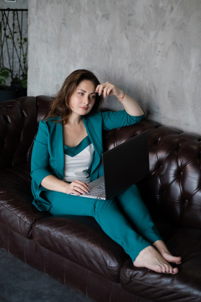 远程工作，远程学习。自信的黑发女人在绿色西装赤脚在皮革沙发与笔记本电脑。家庭办公室。居家办公图片下载