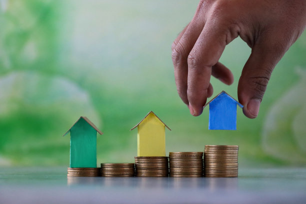 无法辨认的人放置蓝色纸板房子的特写图像，排在多色纸板房子，彩色的海滩小屋硬币堆，重点前景，斑斑驳杂的绿色背景，家庭财务，房地产和假日储蓄概念图片下载
