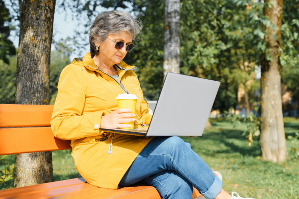 成熟的女人戴着眼镜，穿着休闲，拿着一次性咖啡在笔记本电脑上工作;活跃的退休自由职业者坐在公园的长椅上浏览互联网，远程工作在户外图片下载