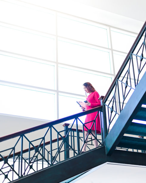 一名女商人穿着鲜艳的裙子，站在灯光明亮的办公大楼的楼梯上，手里拿着文书和手机，同时处理多项任务图片下载