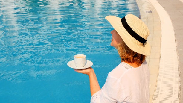 一个50-55岁的女人戴着草帽，在蓝色的游泳池边喝着咖啡，奢侈的早安，一天的开始图片下载