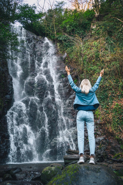一个女人举起双手站在瀑布附近的一块岩石上。一个年轻的女人在山中旅行，在森林中散步，在大自然中放松。旅行和漫游的概念，复制空间。垂直图片下载