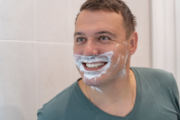一名白人男子剃须时往浴室的镜子里看。一个滑稽的男人对着他的倒影微笑，他的脸被可笑地剃了一半。千禧一代男子打理胡茬或胡须，在沐浴中为面部做清晨美容，sk概念图片下载