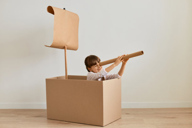 水平拍摄的可爱的小女孩扎着辫子，在装着帆的纸箱里的手工船上玩耍，手里拿着望远镜，眺望远方，探索地平线。图片下载