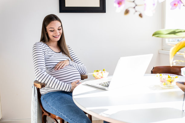 孕妇坐在家里的桌子旁用笔记本电脑进行视频通话。图片下载