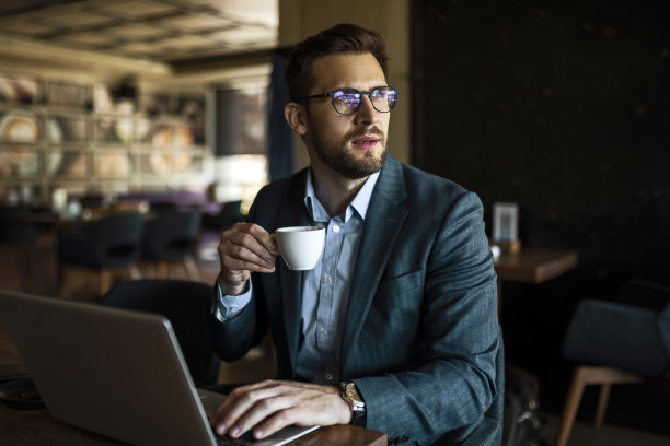商人一边喝咖啡，一边用笔记本电脑工作，看着窗外图片下载