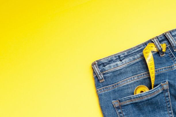 卷尺在蓝色牛仔裤后面的口袋里以黄色为背景，复印空间。概念剪裁，一家服装修理店，正确选择服装尺寸。图片下载