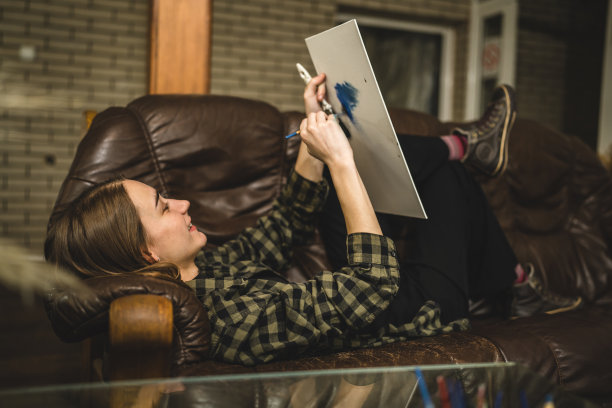 一位年轻美丽的白人女性艺术家坐在床沙发上在画布上绘画，拿着画笔艺术和创意专业或爱好概念真人复制空间侧视图图片下载