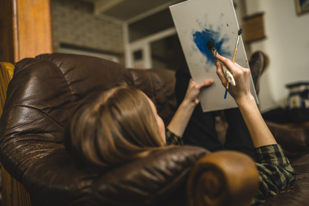 一位年轻美丽的白人女性艺术家坐在床沙发上在画布上绘画，拿着画笔艺术和创意专业或爱好概念真人复制空间侧视图图片下载