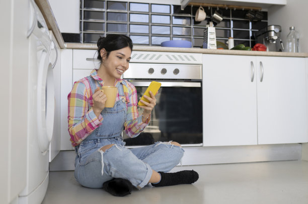 女人在家里的厨房里拿着咖啡和手机坐在地板上——在家放松的概念——图片下载