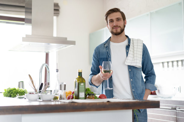 英俊的年轻人穿着便服，站在家里的厨房里喝酒。图片下载