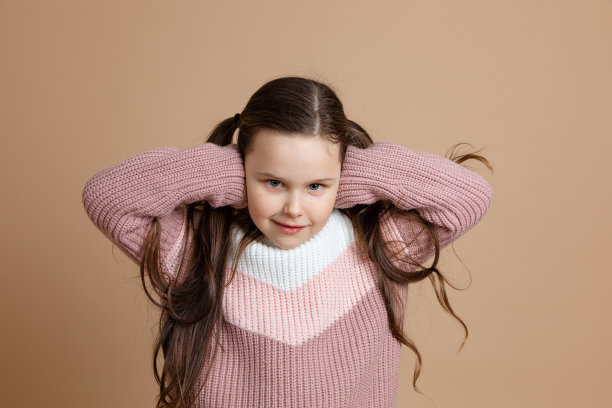 年轻美丽可爱的微笑女孩的肖像，长黑发在白色，粉红色的毛衣站着，用手遮住耳朵，摆姿势。图片下载