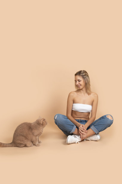 穿着白色内衣和破洞牛仔裤的女人微笑着看着在肤色背景下的桃红色的猫。与宠物玩耍的人。人和动物在一起图片下载
