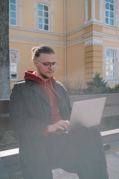 一名年轻的大学生坐在校园的长椅上，通过笔记本电脑在线学习。时尚潮人有经验的自由职业者工作距离笔记本电脑休息图片下载