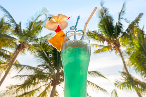 在耀眼的阳光下，在绿色的棕榈树和蓝色的天空的背景下，盛满蓝色饮料的高脚杯。有趣的角度。暑假的概念。图片下载