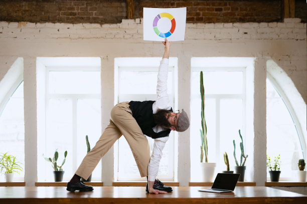 年轻的大胡子，办公室职员，在现代办公室的工作时间，在木桌上用电子设备做瑜伽。商业理念，健康生活方式，运动，爱好图片下载