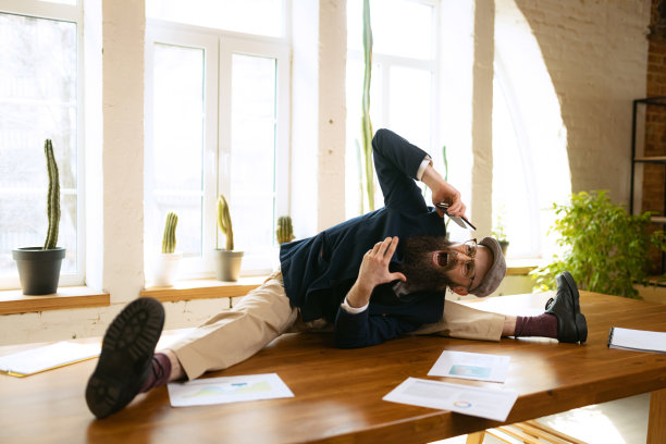 年轻的大胡子，办公室职员，在现代办公室的工作时间，在木桌上用电子设备做瑜伽。商业理念，健康生活方式，运动，爱好图片下载