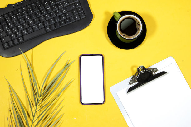 经典的金属剪贴板，空白的白纸和棕榈树叶，黄色背景的黑色键盘的手机，咖啡杯图片下载