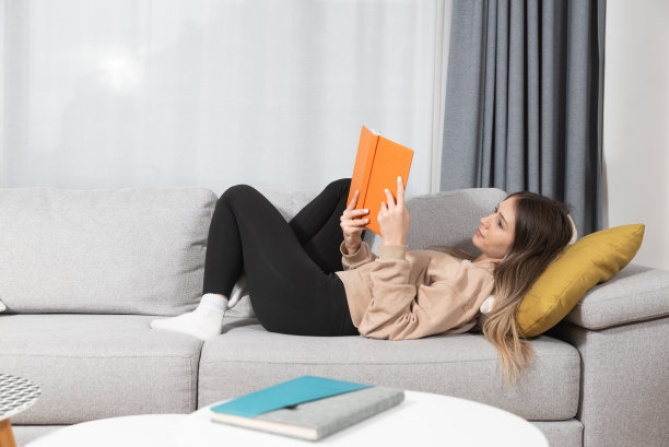 年轻美丽的女人坐在舒适的家里的沙发上看书，享受她的自由时间没有她的丈夫和孩子微笑和幸福的脸。女学生学习和放松。图片下载