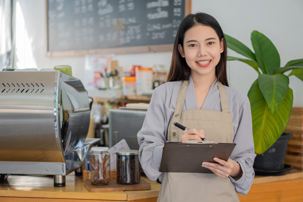 漂亮的亚洲咖啡师，穿着围裙，拿着剪贴板，站在咖啡店柜台前。企业主的创业理念。图片下载