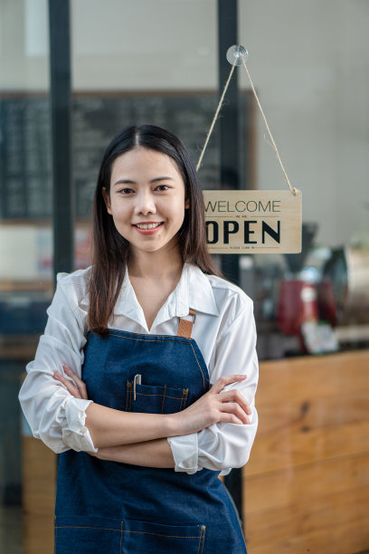 一个穿着围裙的漂亮的亚洲咖啡师站在咖啡馆门口，门口的牌子开着。企业主创业想法图片下载
