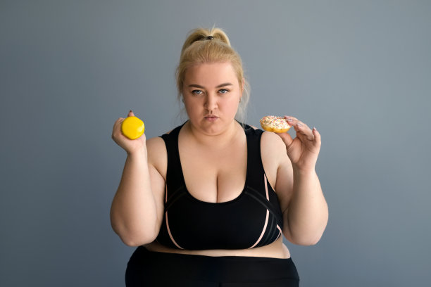 一个年轻的胖女人手里拿着哑铃和百吉饼，一脸饥饿和愤怒。拒绝甜食和面粉有助于减肥。意志力和减肥的欲望图片下载