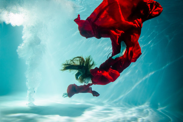 迷人的红发年轻女子穿着红色裙子在水下美丽地游泳。图片下载