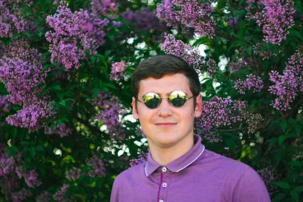 散焦帅哥在圈太阳镜户外肖像。男人微笑着看着镜头。青年男子在紫色衬衣上自然夏天的背景。淡紫色的花。的焦点图片下载