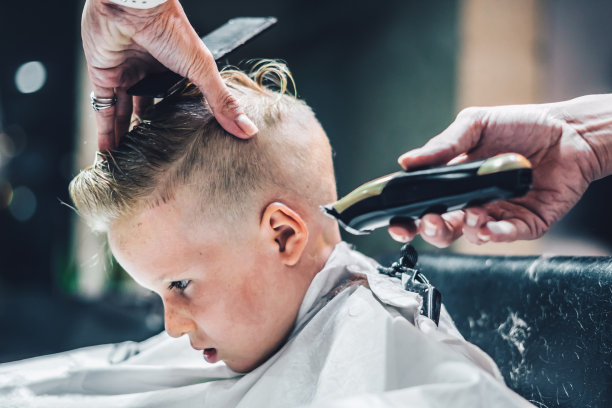 生活照片。理发店的金发小男孩顾客。美发师男士工作时动手梳子工具剪刀修剪白色披肩。现代风格的儿童短发。儿童时尚商务服务图片下载