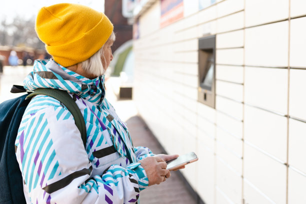 老妇人从包裹柜上拿起包裹。在城市街道的自动邮政终端机附近，一位微笑的女性拿着智能手机。智能快速配送理念。图片下载
