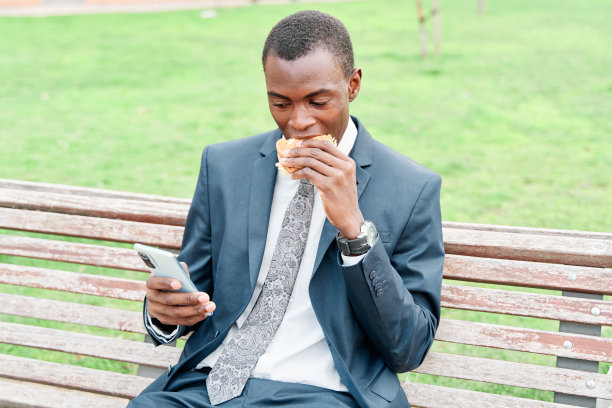 一名非洲裔美国人在户外公园吃汉堡，同时使用智能手机图片下载