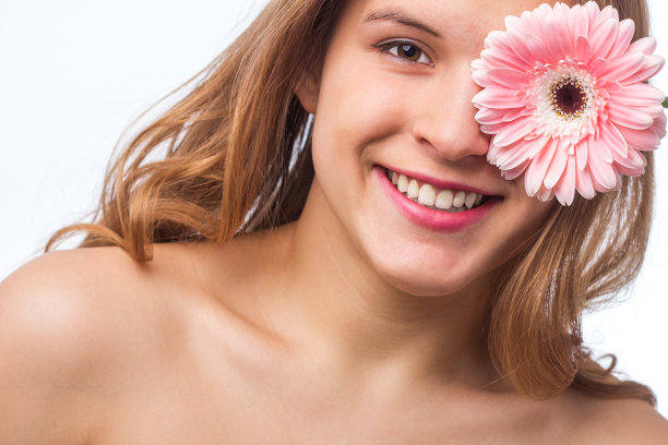 一个快乐的女孩的肖像与新鲜的皮肤拿着一朵花在一只眼睛在白色背景。近距离肖像的美丽的红发卷曲的女人pnik花穿白色t恤。美丽和春天图片下载