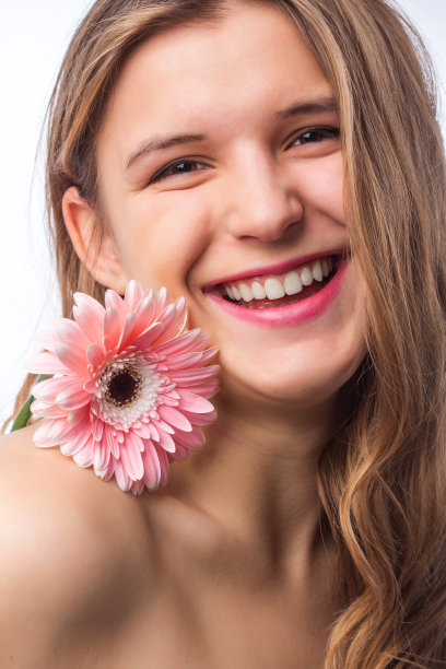 一个快乐的女孩的肖像与新鲜的皮肤拿着一朵花在一只眼睛在白色背景。近距离肖像的美丽的红发卷曲的女人pnik花穿白色t恤。美丽和春天图片下载