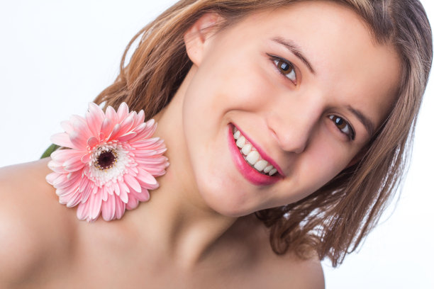 一个快乐的女孩的肖像与新鲜的皮肤拿着一朵花在一只眼睛在白色背景。近距离肖像的美丽的红发卷曲的女人pnik花穿白色t恤。美丽和春天图片下载