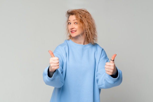 可爱的成熟女人，卷曲的金发，竖起大拇指，ok赞许的手势，推荐的东西，看左边，站在浅灰色背景的时髦的蓝色运动衫图片下载