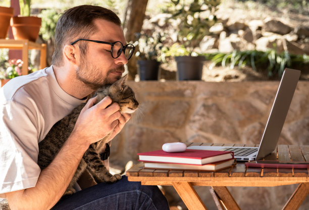 戴眼镜的男人抱着猫，在他的笔记本电脑上工作。宠物的温柔，宠物的爱，在一起和友谊。图片下载