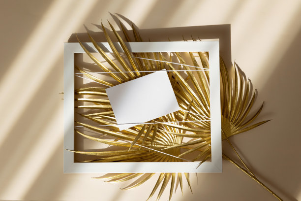 邀请卡模型与金色棕榈叶在白色框架上米色柔和的背景。俯视图，平躺，复制空间。模板空白的白纸模拟为品牌和广告图片下载