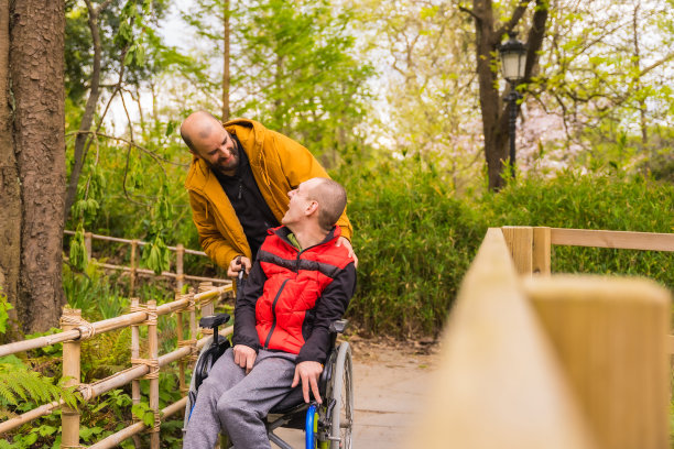 在城市公园里，一个坐在轮椅上的瘫痪青年被朋友推着，彼此微笑着看着对方图片下载
