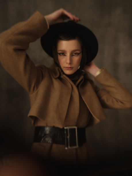 一个美丽的年轻女子在灰色的背景下在工作室里摆姿势。复古外观。秋季或春季外套，黑色帽子和围巾。大眼睛的箭头。图片下载