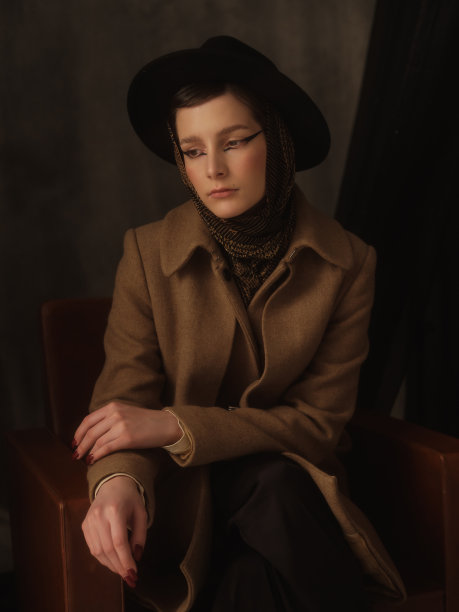 一个美丽的年轻女子在灰色的背景下在工作室里摆姿势。复古外观。秋季或春季外套，黑色帽子和围巾。大眼睛的箭头。图片下载