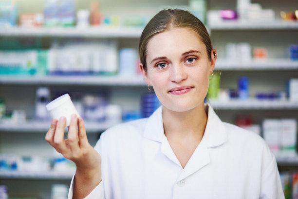 身穿白大褂的年轻女药剂师拿着药瓶，站在装满药品的货架前图片下载