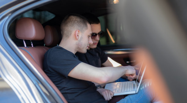 两个朋友坐在车的后座上，看着笔记本电脑，为他们的公路旅行制定计划，寻找他们的目的地。高清库存图片。图片下载